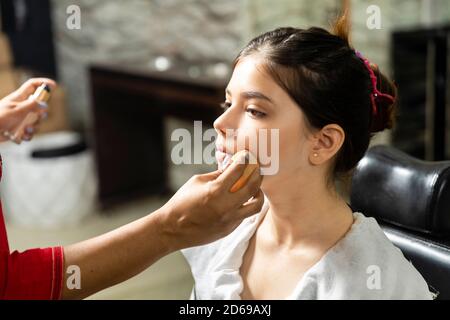 Una bella giovane ragazza indiana ottiene il trucco fatto ad un salone, l'artista del trucco che applica la spugna sulla faccia del modello, trucco e concetto di salone di bellezza. Foto Stock