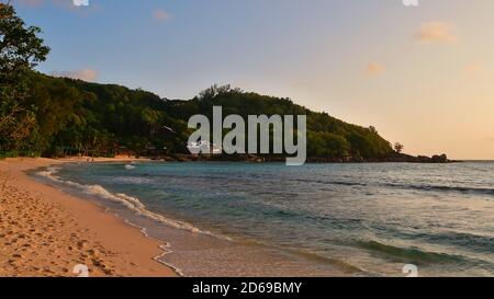 Spiaggia tropicale Anse Takamaka nel sud di Mahe, Seychelles con sabbia, acqua calma, palme, foresta pluviale e alloggio turistico in serata. Foto Stock
