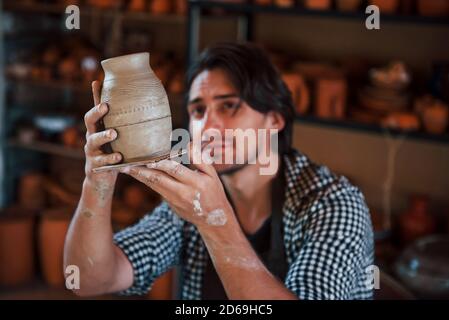 Il giovane ceramista tiene in mano una pentola fresca fatta a mano e guarda al risultato del suo lavoro Foto Stock