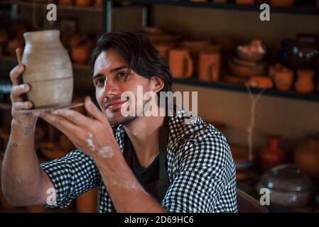 Il giovane ceramista tiene in mano una pentola fresca fatta a mano e guarda al risultato del suo lavoro Foto Stock