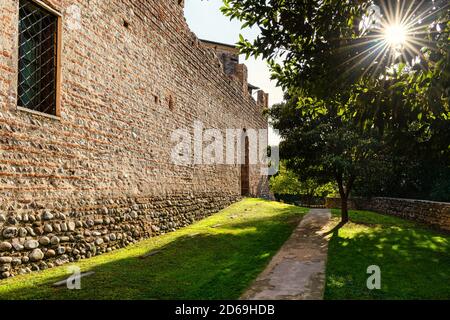 Mura del castello di Bassano del Grappa, Vicenza. I raggi del sole passano attraverso il fogliame degli alberi. Un sentiero nel prato verde Foto Stock