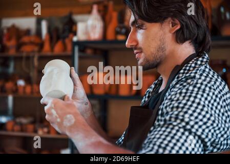 Il giovane ceramista tiene in mano una pentola fresca fatta a mano e guarda al risultato del suo lavoro Foto Stock