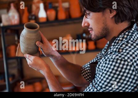 Il giovane ceramista tiene in mano una pentola fresca fatta a mano e guarda al risultato del suo lavoro Foto Stock