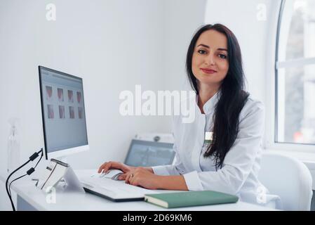 Bruna femmina medico si siede in moderno ufficio da computer e. guarda la fotocamera Foto Stock