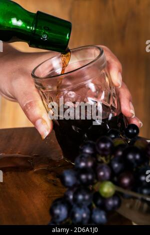 Versando il vino in un bicchiere di cristallo da una bottiglia verde, una mano caucasica tiene il calice nel processo, ci sono piccole uve mature in un bl Foto Stock