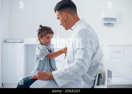 Insegna alla ragazza come usare stetoscopio. Il pediatra giovane lavora con la piccola visitatrice femminile nella clinica Foto Stock