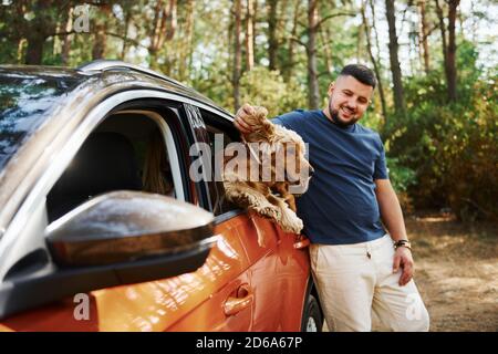 L'uomo si trova accanto all'auto nella foresta. Cute cane in veicolo guarda attraverso il finestrino Foto Stock