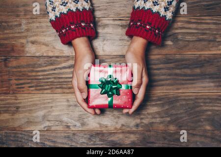 Mani femminili con un maglione invernale che tiene un regalo di Natale su un rustico tavolo di legno. Dare o ricevere concetto. Vista dall'alto Foto Stock