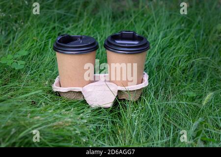 Due tazze di carta marrone con coperchi neri in un supporto contro uno sfondo di erba. Ci sono due bicchieri di caffè sul prato. Tazze di carta da tè con plasti Foto Stock