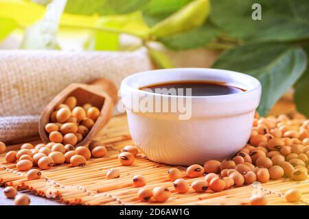 Salsa di soia con fagioli di soia sparsi su un tovagliolo di bambù sullo sfondo del burlap e ramificazione giovani baccelli verdi di soia, closeup con fo selettivo Foto Stock