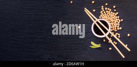 Salsa di soia in una ciotola con chopsticks di bambù e fagioli di soia sparsi sulla superficie nera della pietra di ardesia, striscione, vista dall'alto, closeup con spazio per Foto Stock