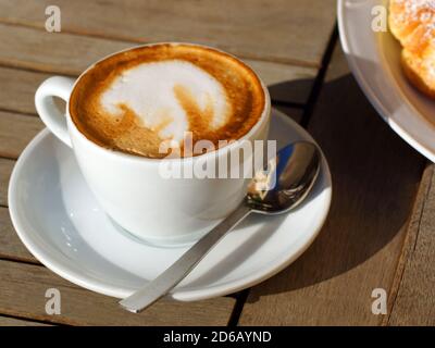 Deliziosi hot cappuccino italiano sorge in una tazza con un piattino su un tavolo di legno. Sullo sfondo di un croissant fresco su una piastra. Foto Stock