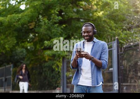 Passeggiate per la strada, relax, tempo libero. Giovane uomo afro-americano in cuffia che ascolta la musica sullo smartphone utilizzando l'app musicale. Ritratto di sorridere Foto Stock