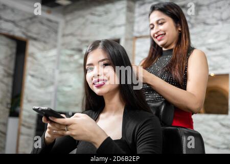 Donna parrucchiere in piedi e facendo acconciatura a bella giovane donna in salone di bellezza, sorridente giovane ragazza che tiene il suo telefono cellulare. Foto Stock