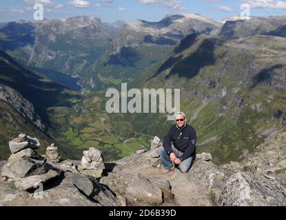 Squatting Man al Dalbinbba Lookout con le grandi montagne di Geirangerfjord in background in UN giorno estivo soleggiato con Un cielo blu chiaro e alcuni Foto Stock