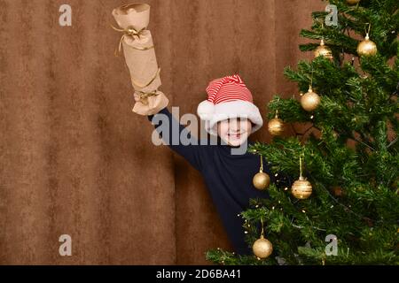 Un ragazzo con un flapper fatto in casa, sbirciando da dietro l'albero di Natale. Sorride allegro. Foto Stock