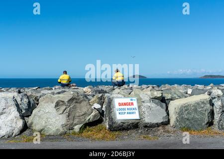 Mackay, Queensland, Australia - 2020 giugno: Due lavoratori che prendono una pausa con le loro canne da pesca sul muro di roccia del porto, ignorando il segno di tenere lontano Foto Stock