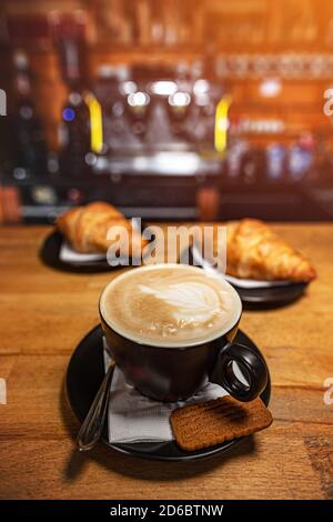 Tazza di caffè e croissant su tavolo di legno con sfocatura caffetteria come sfondo Foto Stock