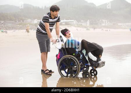 Giovane uomo caucasico asiatico biraciale che spinge il fratello minore disabile dentro sedia a rotelle sulla spiaggia Foto Stock