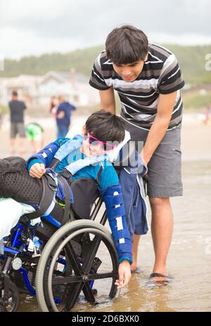 Giovane uomo caucasico asiatico biraciale che spinge il fratello minore disabile dentro sedia a rotelle sulla spiaggia Foto Stock