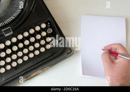 una mano maschile che scrive su una carta a scacchi bianca con una penna rossa accanto a una vecchia macchina da scrivere nera distrutta. Concetto di creatività e narrazione Foto Stock