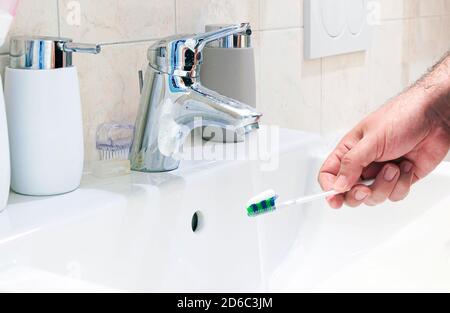 Una mano maschile che tiene uno spazzolino da denti con dentifricio in un lavello in ceramica bianca di un bagno. Pulizia dentale e igiene personale. Routine mattutina Foto Stock