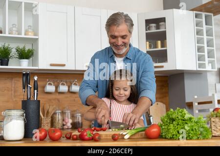 Carino bambina e il suo bel papà stanno tagliando verdure e sorridendo mentre cucinando in cucina a casa. Foto Stock