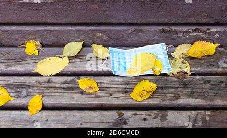 maschera sanitaria usata coperta da foglie gialle cadenti vecchia panca di legno nel parco della città il giorno d'autunno Foto Stock