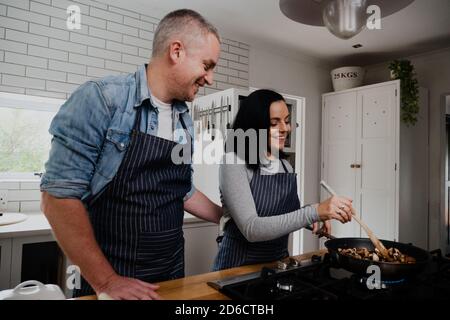 Felice coppia che indossa grembiuli facendo una sana cena per i bambini in piedi in cucina moderna. Foto Stock