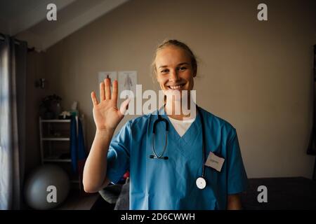 Infermiera caucasica che agitava e salutava mentre era in videochiamata in piedi indossare scrub in clinica Foto Stock
