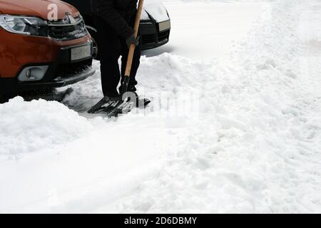 Pala in mano. L'uomo con pala libera la neve intorno all'auto nel parcheggio in inverno dopo la nevicata. Problemi invernali dei conducenti di auto. Foto Stock