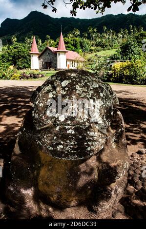 Antica statua in pietra sulla spiaggia di Nuku Hiva, chiesa distante sullo sfondo Foto Stock