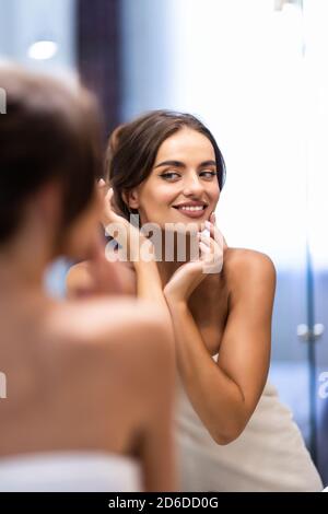 Bella sorridente giovane donna in accappatoio e asciugamano sulla testa guardando lo specchio nella stanza da bagno Foto Stock