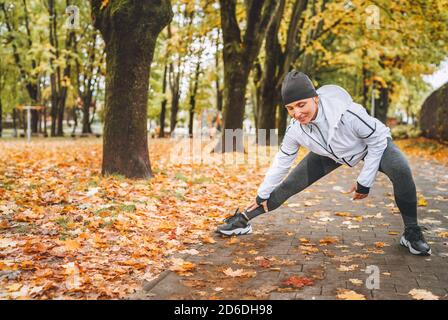 Donna atletica in forma che fa esercizio prima di fare jogging nel parco cittadino autunnale sul parco giochi per bambini. Fitness femminile di mezza età che allunga le gambe mentre Foto Stock