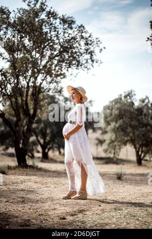 Donna incinta che indossa cappello e abito bianco tra gli alberi di sughero Foto Stock