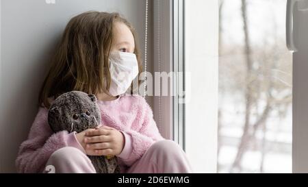 COVID-19 concetto di coronavirus, ragazzino triste in maschera medica guarda fuori finestra. Il bambino pensivo si siede sul davanzale a casa in autunno o in inverno. Bambina con Foto Stock