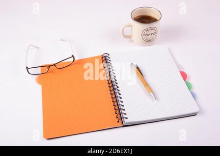 Taccuino arancione aperto con una penna, bicchieri e una tazza di caffè su sfondo bianco Foto Stock