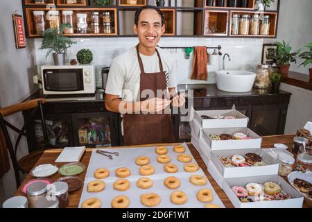 Sorridente maschio asiatico che indossa un grembiule utilizzando uno smartphone in piedi ciambelle anteriori su un tavolo in cucina Foto Stock