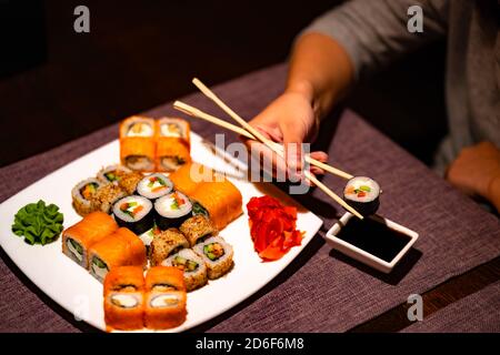 Persona che mangia sushi con chopsticks nel ristorante. Crop persona anonima con chopsticks mangiare vari tipi di sushi panini serviti con wasabi e. Foto Stock