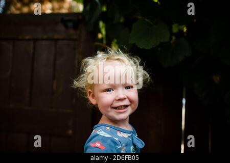 Biondo ragazzo di due anni sorridente per la fotocamera in piedi Iarda Foto Stock
