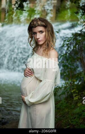 Giovane bella ragazza incinta in un vestito bianco e corona si erge sullo sfondo di una cascata Foto Stock