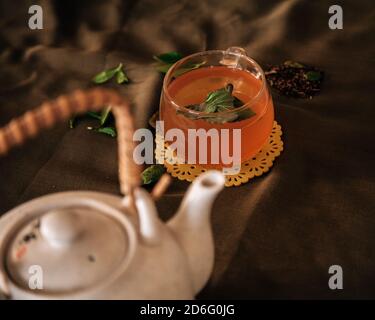 Il bicchiere di tè verde e le foglie di erbe naturali accanto alla ceramica bianca Tradizionale piatto da tè asiatico su sfondo scuro Foto Stock