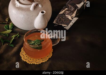 Il bicchiere di tè verde e le foglie di erbe naturali accanto alla ceramica bianca Tradizionale piatto da tè asiatico su sfondo scuro Foto Stock