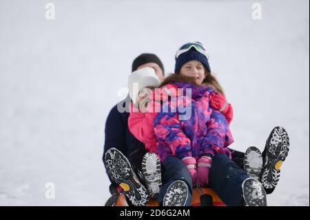 felice uomo di famiglia con due ragazze che cavalcano sul tubo sopra neve bianca nella località invernale Foto Stock