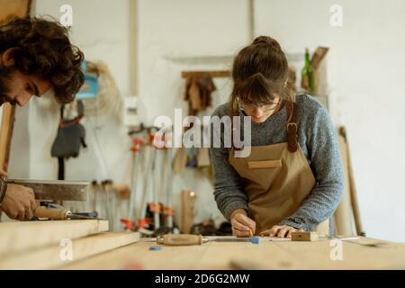 Una giovane coppia di falegnami che lavorano insieme in una piccola officina di carpenteria che progetta una nuova parte domestica di mobilia che si sta costruendo fuori del legno. Giovane entrpr Foto Stock