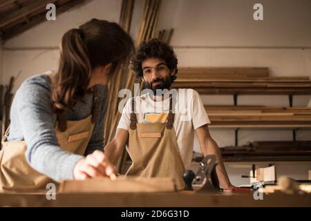 Una giovane coppia di falegnami che lavorano insieme in una piccola officina di carpenteria che progetta una nuova parte domestica di mobilia che si sta costruendo fuori del legno. Giovane entrpr Foto Stock