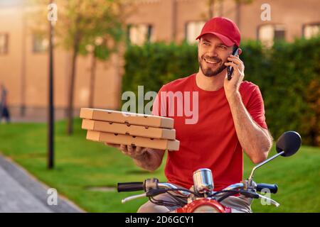 Felice giovane corriero caucasico maschio con pizza che parla al telefono mentre si siede su scooter all'aperto. Concetto di consegna degli alimenti Foto Stock