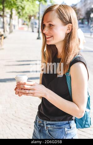 ritratto verticale di una giovane bella donna caucasica rossa in piedi in strada con una tazza di caffè da portare via e sorridendo Foto Stock