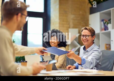 Uomo allegro che dà una cartella con i documenti al suo collega e sorridente mentre lavora insieme nell'ufficio moderno. Lavoro di squadra e collaborazione, concetto di business people Foto Stock