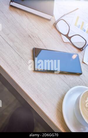 Lavoro in caffetteria. Vista dall'alto di uno smartphone, di una tazza di caffè e di occhiali sul tavolo in legno, a scatto verticale Foto Stock